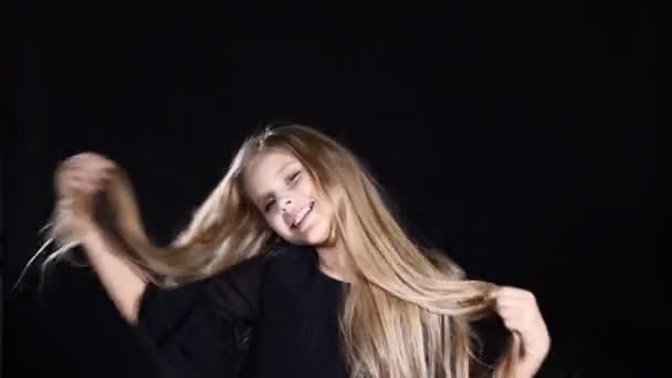 Portrait of adorable girl posing looking at camera. Smiling and touching her hair. Blond pretty teen girl with long hair on black background. - Footage, Video