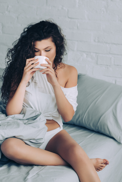 attractive curly woman drinking coffee in bed during morning time at home - Photo, Image