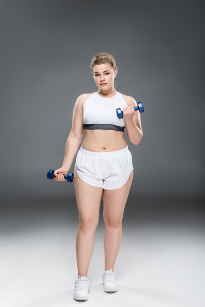 young oversize woman in sportswear exercising with dumbbells and looking at camera on grey  - Foto, Imagem