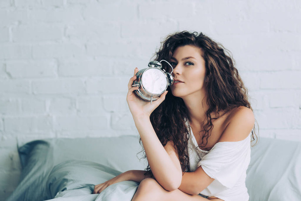 cheerful curly young woman sitting on bed with alarm clock at home - 写真・画像