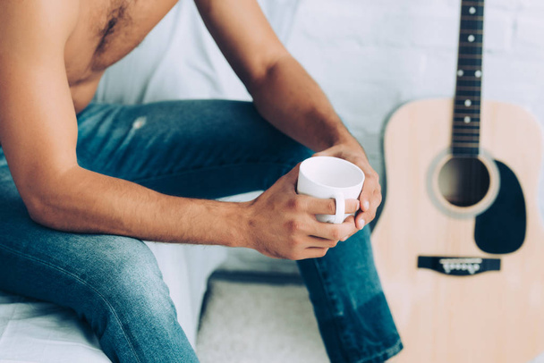 gedeeltelijke weergave van shirtless man in spijkerbroek koffiekopje drinken tijdens ochtend tijd in slaapkamer thuis - Foto, afbeelding