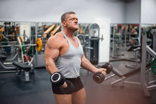 Big athletic man trains in the gym. Training biceps with a dumbbells - Zdjęcie, obraz