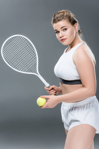 young oversize woman with tennis racket and ball looking at camera on grey - Φωτογραφία, εικόνα