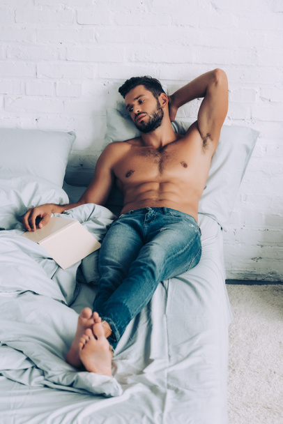 young shirtless muscular man sitting with book on bed at home - Foto, Imagem