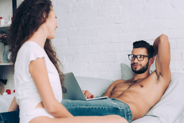 cheerful shirtless male freelancer in eyeglasses working on laptop and talking to girlfriend in bedroom at home - Fotoğraf, Görsel