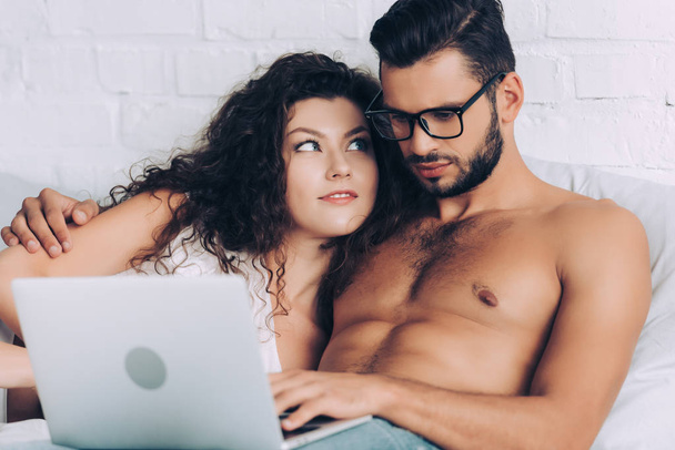 joven sin camisa freelancer masculino en gafas que trabajan en el ordenador portátil y abrazar a la novia en el dormitorio en casa
 - Foto, imagen