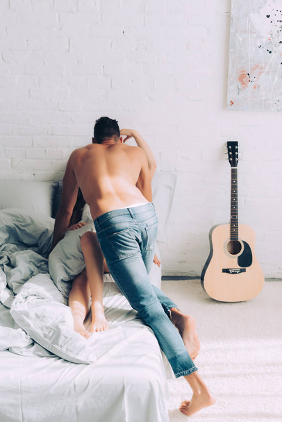 zadní pohled na košili svalnatý muž v džínách s přítelkyní na posteli doma - Fotografie, Obrázek