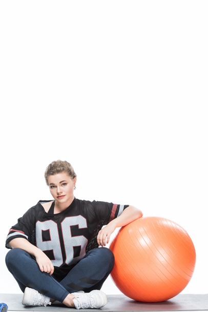giovane donna in sovrappeso in abbigliamento sportivo appoggiata a palla in forma e seduta su tappetino yoga isolato su bianco
  - Foto, immagini