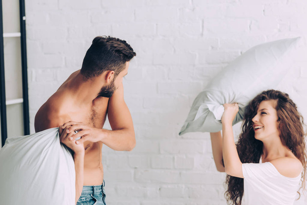 selective focus of smiling young couple having fun and fighting by pillows in bedroom at home - Photo, Image