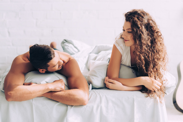 χαρούμενο ζευγάρι μιλάμε και τοποθέτηση στο κρεβάτι με τα μαξιλάρια πρωί στο σπίτι - Φωτογραφία, εικόνα