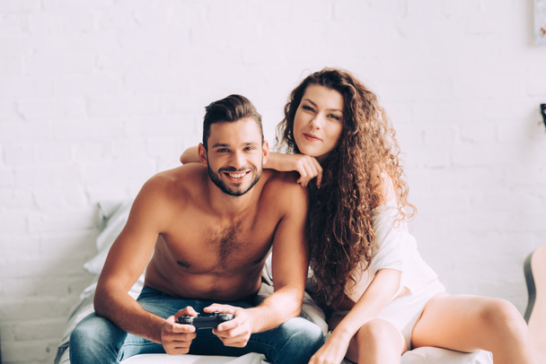счастливый мужчина без рубашки играет в видеоигру с джойстиком в то время как его кудрявая подруга сидит рядом на кровати дома
 - Фото, изображение