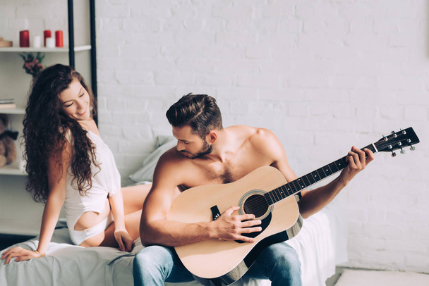 м'язистий без сорочки чоловік грає на акустичній гітарі, а його дівчина сидить поруч у спальні вдома
 - Фото, зображення