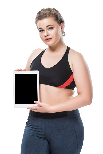 giovane donna in sovrappeso in abbigliamento sportivo in possesso di tablet digitale e guardando la fotocamera isolata su bianco
  - Foto, immagini