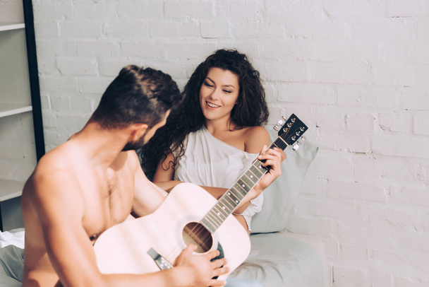 Mann spielt auf Akustikgitarre, während seine glückliche Freundin zu Hause im Schlafzimmer sitzt - Foto, Bild