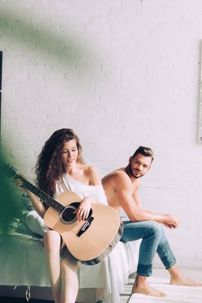 selective focus of curly woman playing on acoustic guitar while her boyfriend sitting near on bed at home - Φωτογραφία, εικόνα