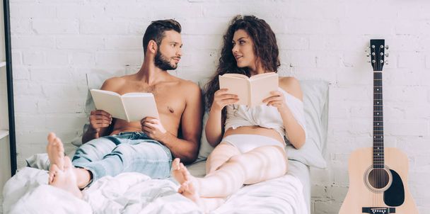 happy young couple sitting with books and looking at each other on bed at home - Foto, afbeelding