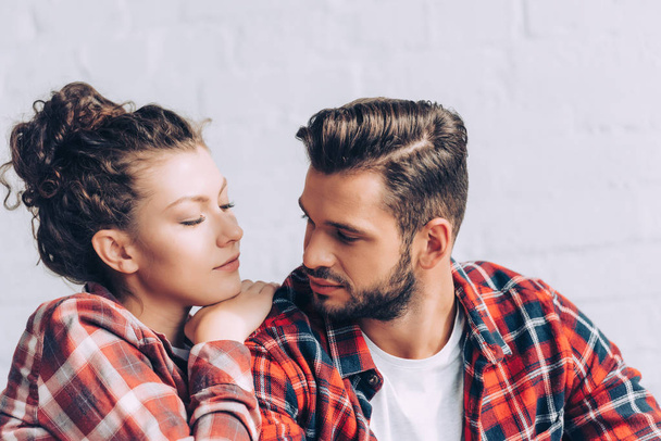 pretty young woman in checkered shirt embracing boyfriend at home - Фото, изображение