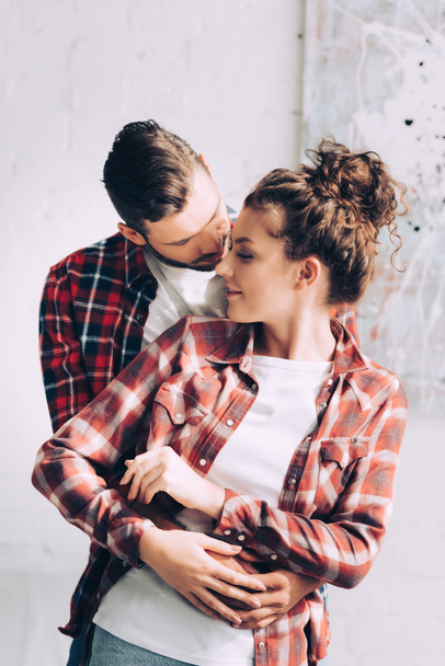 young handsome man in checkered shirt embracing smiling girlfriend at home - Fotó, kép