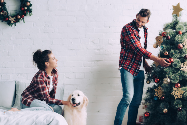 vista lateral de niña acariciando golden retriever mientras su novio decorando el árbol de Navidad en casa
 - Foto, Imagen