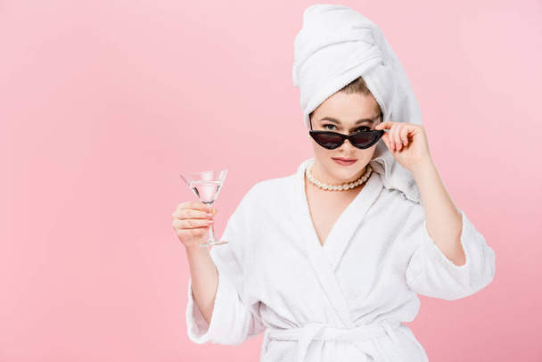 atraktivní oversize žena v županu, sluneční brýle a ručník na hlavě drží sklo a při pohledu na fotoaparát izolované na růžové - Fotografie, Obrázek