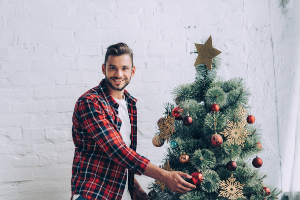 happy young man decorating christmas tree at home - Zdjęcie, obraz