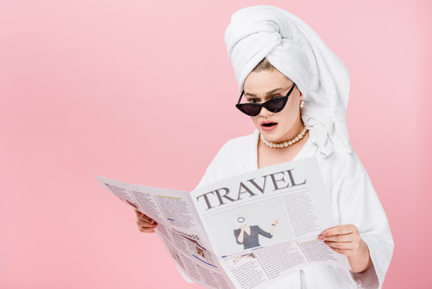 σοκαρισμένος νέους μέγεθος συν γυναίκα στο μπουρνούζι, γυαλιά ηλίου και πετσέτα στο κεφάλι διαβάζοντας εφημερίδα ταξιδιού που απομονώνονται σε ροζ - Φωτογραφία, εικόνα