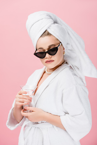 young oversize woman in bathrobe, sunglasses and towel on head holding glass isolated on pink - Photo, Image