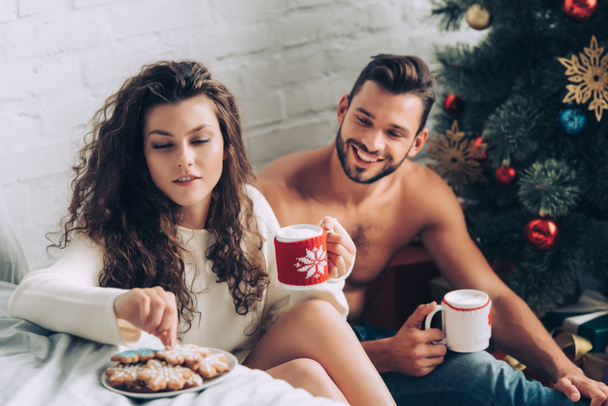 young curly woman with coffee cup taking ginger cookie while her boyfriend sitting near christmas tree at home - Foto, imagen
