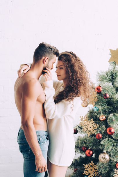 веселая молодая женщина обнимает бойфренда без рубашки возле рождественской елки дома
 - Фото, изображение