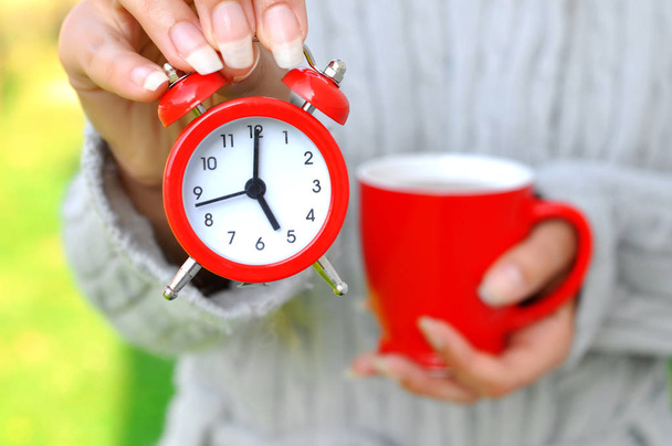 Žena drží červené hodinky a červená šálek teplého nápoje. To ukazuje, že je čas na přestávku. Práce je u konce a je čas pro radost útulný domov. Selektivní fokus - Fotografie, Obrázek
