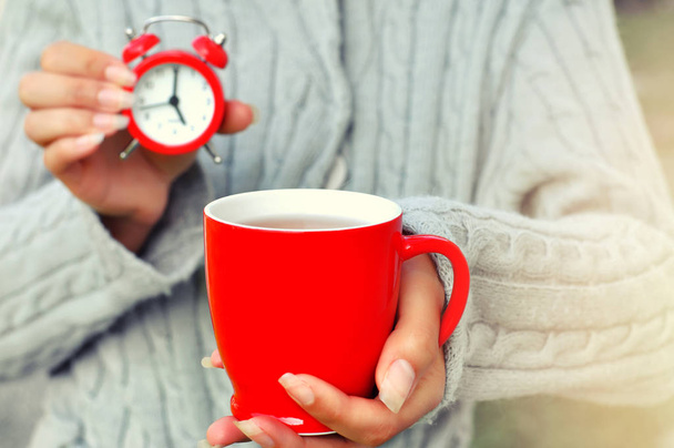 Žena drží červené hodinky a červená šálek teplého nápoje. To ukazuje, že je čas na přestávku. Práce je u konce a je čas pro radost útulný domov. Selektivní fokus - Fotografie, Obrázek