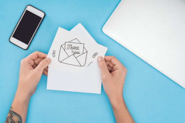 pessoa cortada segurando papel com obrigado lettering saindo do envelope e smartphone isolado no fundo azul
 - Foto, Imagem