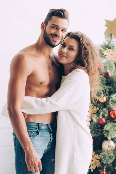 σγουρά χαμογελαστή γυναίκα αγκαλιάζει shirtless μυϊκή φίλο κοντά στο χριστουγεννιάτικο δέντρο στο σπίτι - Φωτογραφία, εικόνα