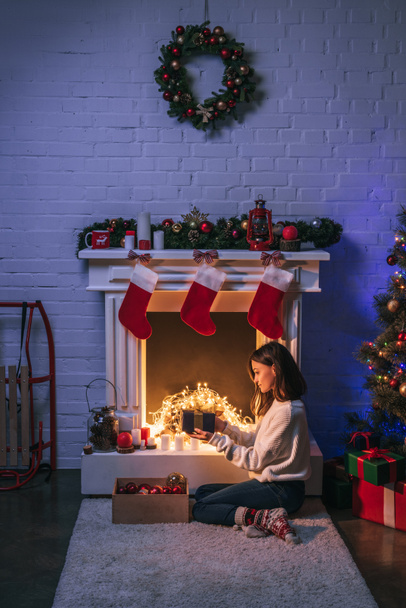 beautiful woman holding gift and sitting near christmas decorated fireplace and Christmas tree - Photo, Image