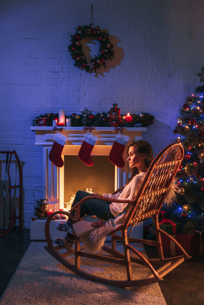 femme heureuse assise dans le fauteuil à bascule près de cheminée décorée et arbre de Noël
 - Photo, image