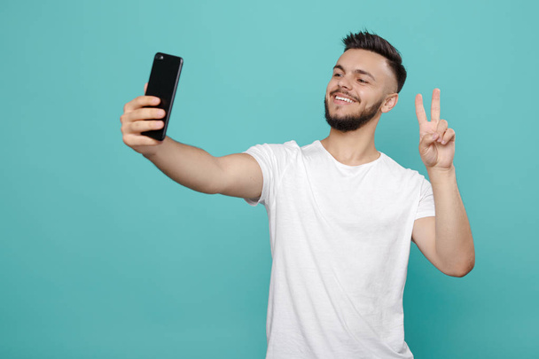 Handsome young bearded hipster in white t-shirt using phone and taking selfie with bright smile. - Fotografie, Obrázek