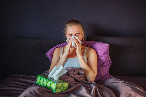 Больной и больной молодой женщины чихающей в белую салфетку. Она сидит на кровати и покрыта шелковым одеялом. У молодой женщины на кровати зеленая коробка с салфетками.
. - Фото, изображение