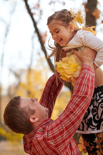 Ο πατέρας πετάει ψηλά κόρη φθινόπωρο πάρκο της πόλης. Αυτοί που θέτουν, χαμογελώντας, παίζοντας. Φωτεινά κίτρινα δέντρα. - Φωτογραφία, εικόνα