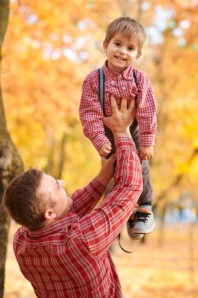 Father throws the boy high up. Father and son are playing and having fun in autumn city park. They posing, smiling, playing. Bright yellow trees. - 写真・画像