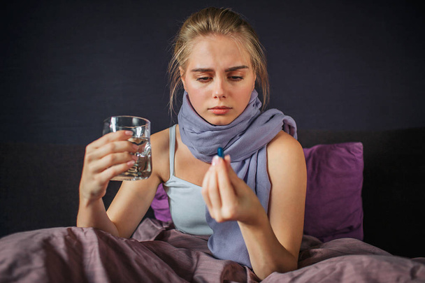 Concentrated young woman sits on bed and looks at medicine she holds with fingers. There is glass of water in another hand. - Foto, afbeelding