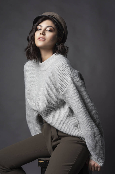 Beautiful brunette girl wearing casual style knitted sweater, brown pants and a hat, sits on a stool on a gray background. Natural nude makeup. Fashion, advertising and commercial design. Copy space. - Fotó, kép