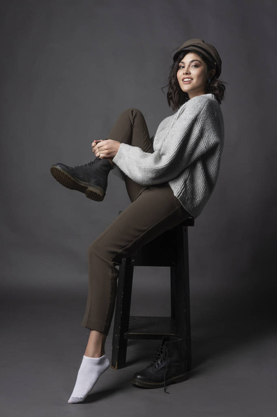 Beautiful smiling brunette girl wearing a casual style sweater, pants and cap puts a shoe on her leg on a gray background. Natural nude makeup. Fashion, advertising and commercial design. Copy space. - Photo, image