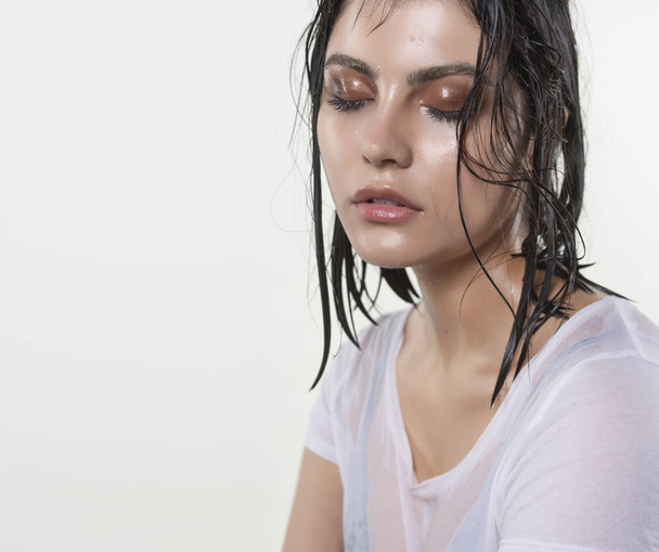 Beautiful wet brunette girl with water drops running down her face, wearing a white translucent T-shirt, through which a black bra shines through. Isolated on white background. Natural makeup. - Foto, afbeelding