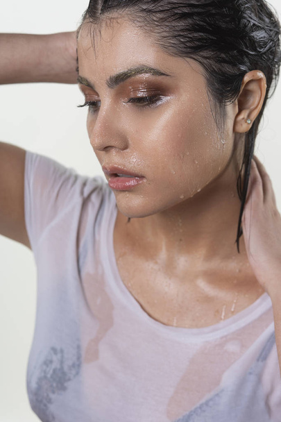 Beautiful wet brunette girl with water drops running down her face, wearing a white translucent T-shirt, through which a black bra shines through. Isolated on white background. Natural makeup. - Photo, Image