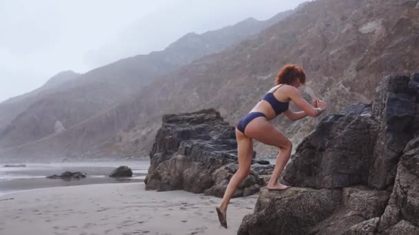 Girl athlete performs squats on one leg standing on a rock on the beach with black sand during training on a beautiful beach. Healthy lifestyle while traveling - Footage, Video