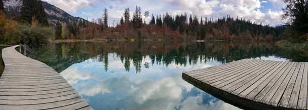 Панорамный пейзаж осеннего цветного леса и листвы окружают идиллическое горное озеро в Альпах Швейцарии поздним осенним днем с отражениями в воде с деревянной набережной на переднем плане
 - Фото, изображение