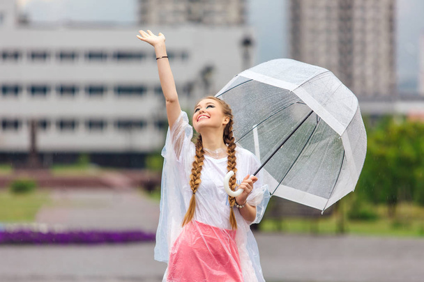 junges hübsches Mädchen mit zwei Zöpfen in gelben Stiefeln und mit transparentem Regenschirm steht in der Nähe des Brunnens. Regentag in der Stadt. - Foto, Bild