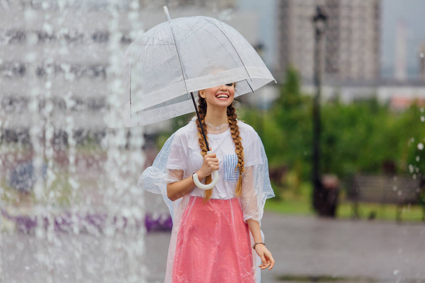 Genç güzel kız sarı çizmeli iki örgüler ve şeffaf şemsiye ile Çeşme duruyor. Şehirde yağmurlu gün. - Fotoğraf, Görsel