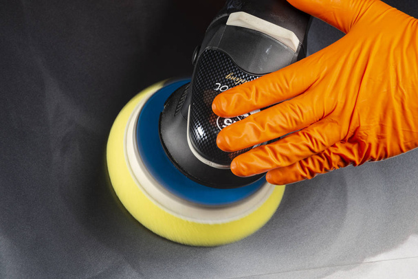 The man is polishing dark color car with polish machine. Close-up view of a hands in orange gloves holding buffer - Фото, зображення