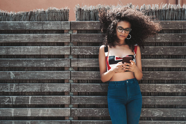 Charming young biracial female leaning against a wooden hedge with wattle fence inside and checking incoming messages on her smartphone, with a copy space area on the left for your text or logo - Fotó, kép